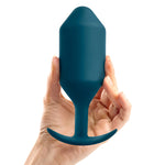 B-Vibe Snug Plug 6 (XXXL). A light skinned hand holding a large, marine blue, cylindrical butt plug with a slightly pointed tip, thick neck, and curved flared base