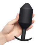 B-Vibe Snug Plug 7 (XXXXL). A light skinned hand holding a large black cylindrical butt plug with a slightly pointed tip, thick neck, and curved flared base