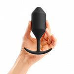 B-Vibe Snug Plug 4 (XL). A light skinned hand holding a large black cylindrical butt plug with a slightly pointed tip, narrow neck, and curved flared base