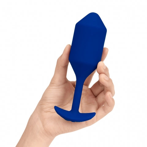 B-Vibe Snug Plug Vibrating XL. A light skinned hand holding a large navy blue cylindrical butt plug with a slightly pointed tip, long thin neck, and curved flared base