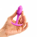 B-Vibe Snug Plug 1 (Small). A light skinned hand holding a small pink cylindrical butt plug with a slightly pointed tip, long narrow neck, and curved flared base